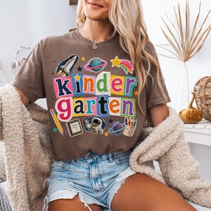 May include: A brown short-sleeve t-shirt featuring the word kindergarten in colorful, multi-patterned block letters. The design includes space-themed graphics like a rocket, planets, an astronaut, a pencil, a notebook, and crayons.