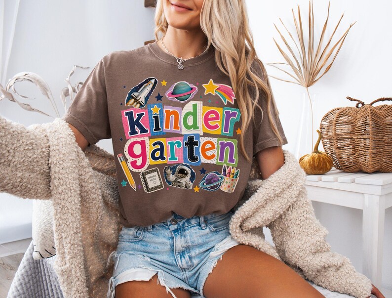 May include: A brown short-sleeve t-shirt featuring the word kindergarten in colorful, multi-patterned block letters. The design includes space-themed graphics like a rocket, planets, an astronaut, a pencil, a notebook, and crayons.