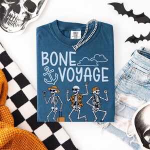 May include: A blue t-shirt with the words "BONE VOYAGE" and a graphic of dancing skeletons. The skeletons are wearing sunglasses, hats, and Hawaiian shirts. A silver chain necklace is on top of the shirt. Halloween-themed decor surrounds the shirt.