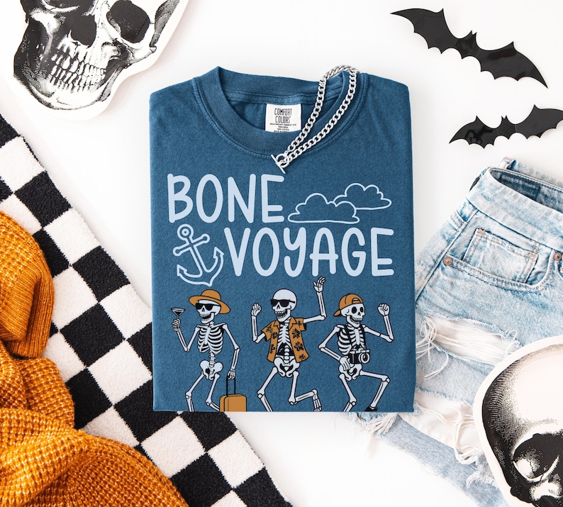May include: A blue t-shirt with the words "BONE VOYAGE" and a graphic of dancing skeletons. The skeletons are wearing sunglasses, hats, and Hawaiian shirts. A silver chain necklace is on top of the shirt. Halloween-themed decor surrounds the shirt.