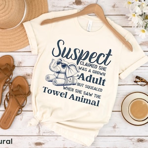 May include: A cream-coloured t-shirt with a blue graphic of an elephant with a towel on its head. The text on the shirt reads "Suspect Claimed She Was A Grown Adult But Squealed When She Saw The Towel Animal".