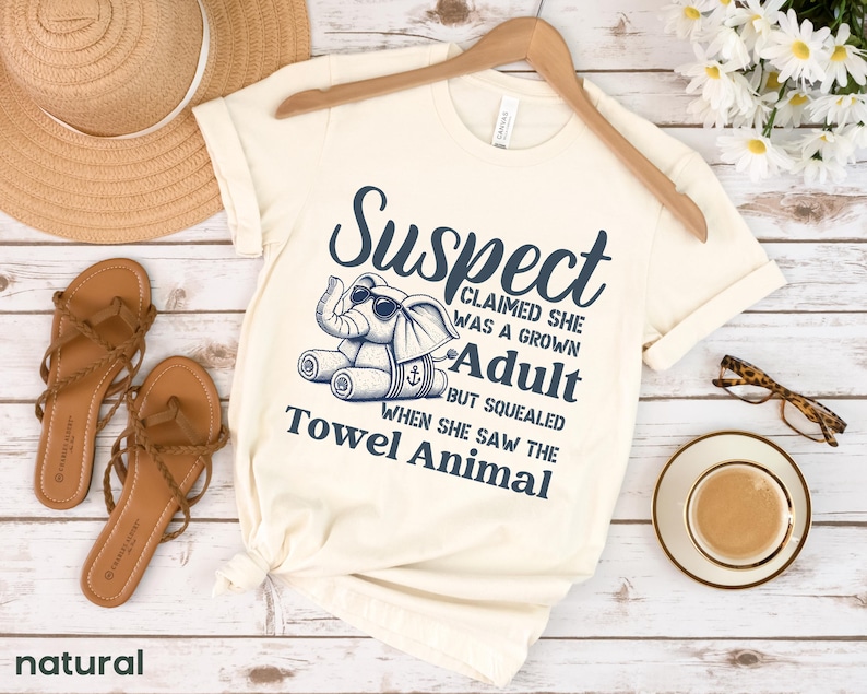 May include: A cream-coloured t-shirt with a blue graphic of an elephant with a towel on its head. The text on the shirt reads "Suspect Claimed She Was A Grown Adult But Squealed When She Saw The Towel Animal".