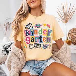 May include: A butter yellow short-sleeve t-shirt featuring the word Kindergarten in colorful, blocky, cutout-style letters. The graphic includes space-themed illustrations like a rocket, planets, an astronaut, a pencil, and a notebook.