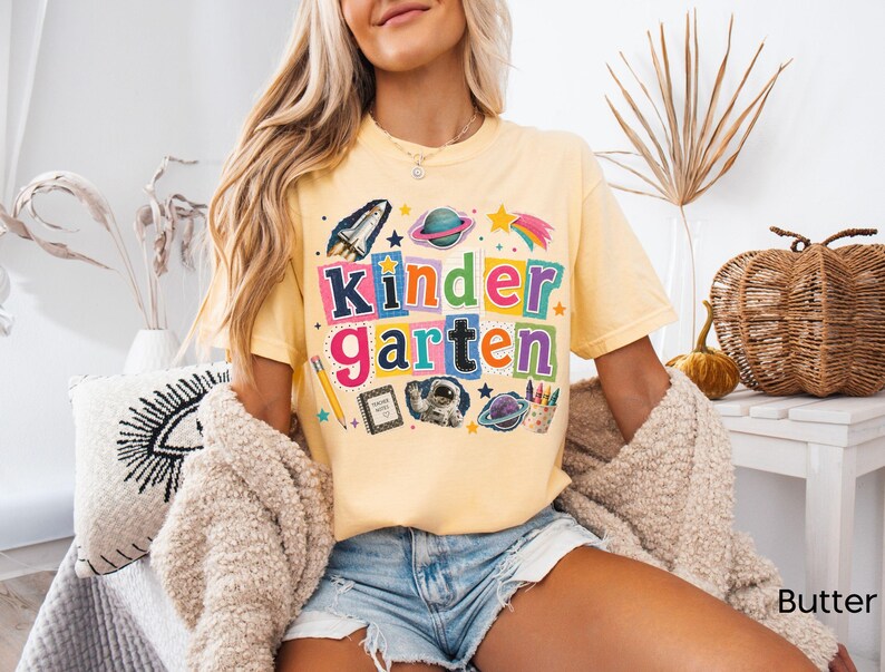 May include: A butter yellow short-sleeve t-shirt featuring the word Kindergarten in colorful, blocky, cutout-style letters. The graphic includes space-themed illustrations like a rocket, planets, an astronaut, a pencil, and a notebook.
