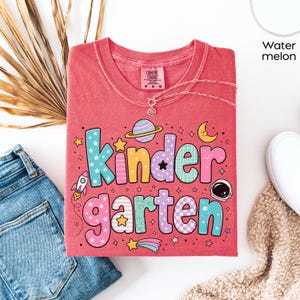 May include: A watermelon-colored t-shirt featuring the word Kindergarten in large, multi-colored, patterned bubble letters. The design includes space-themed graphics such as a planet, a rocket, a crescent moon, stars, and an astronaut helmet.