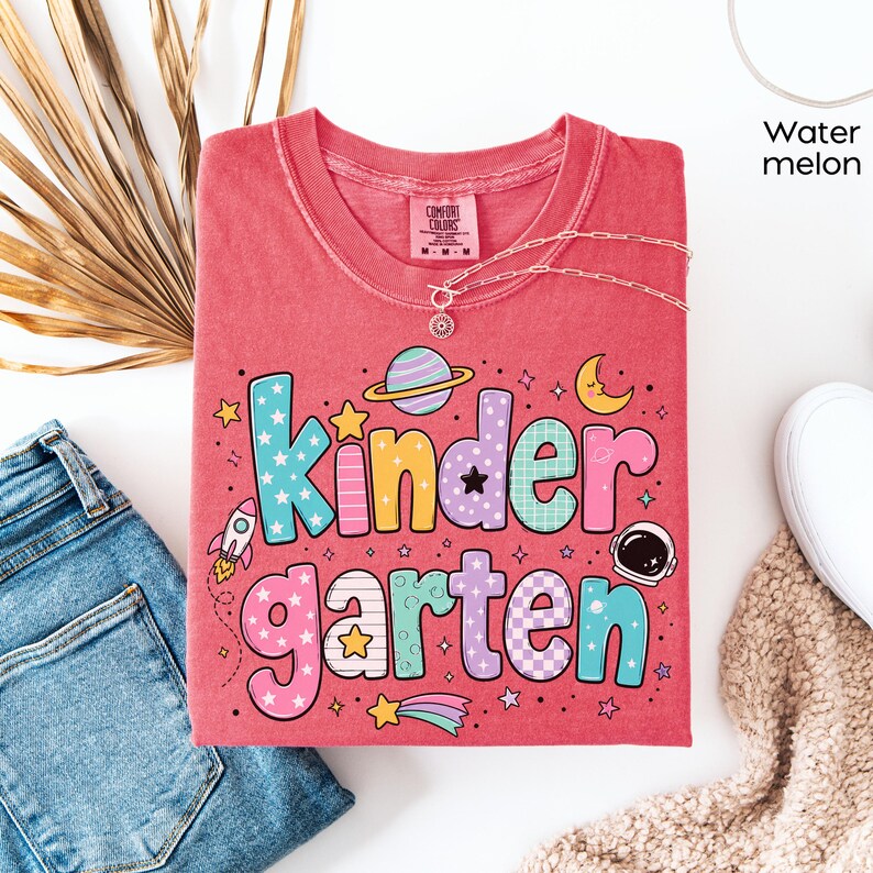 May include: A watermelon-colored t-shirt featuring the word Kindergarten in large, multi-colored, patterned bubble letters. The design includes space-themed graphics such as a planet, a rocket, a crescent moon, stars, and an astronaut helmet.