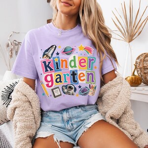 May include: A light purple short-sleeve t-shirt featuring a colorful graphic with the text "kindergarten" in block letters. The design includes illustrations of a space shuttle, planets, a shooting star, a pencil, a notebook, an astronaut, and crayons.