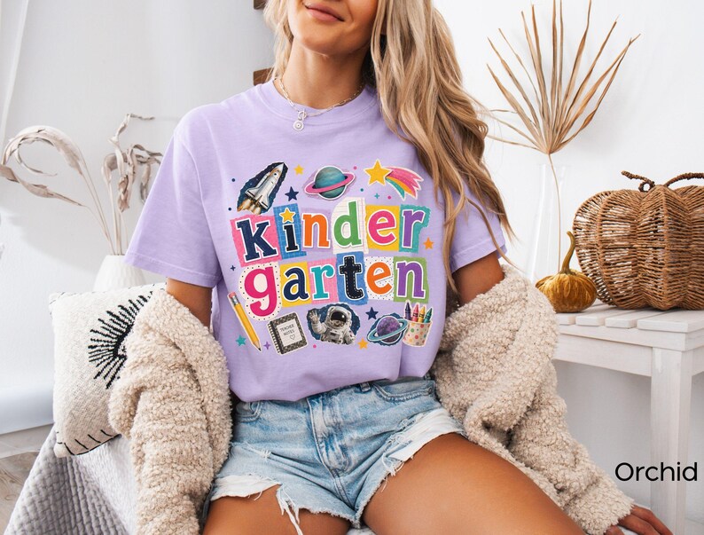 May include: A light purple short-sleeve t-shirt featuring a colorful graphic with the text "kindergarten" in block letters. The design includes illustrations of a space shuttle, planets, a shooting star, a pencil, a notebook, an astronaut, and crayons.