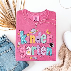 May include: A pink short-sleeve t-shirt featuring the text "Kindergarten" in large, multi-colored bubble letters. The design includes space-themed graphics such as a rocket, a planet with rings, an astronaut helmet, stars, and a crescent moon.