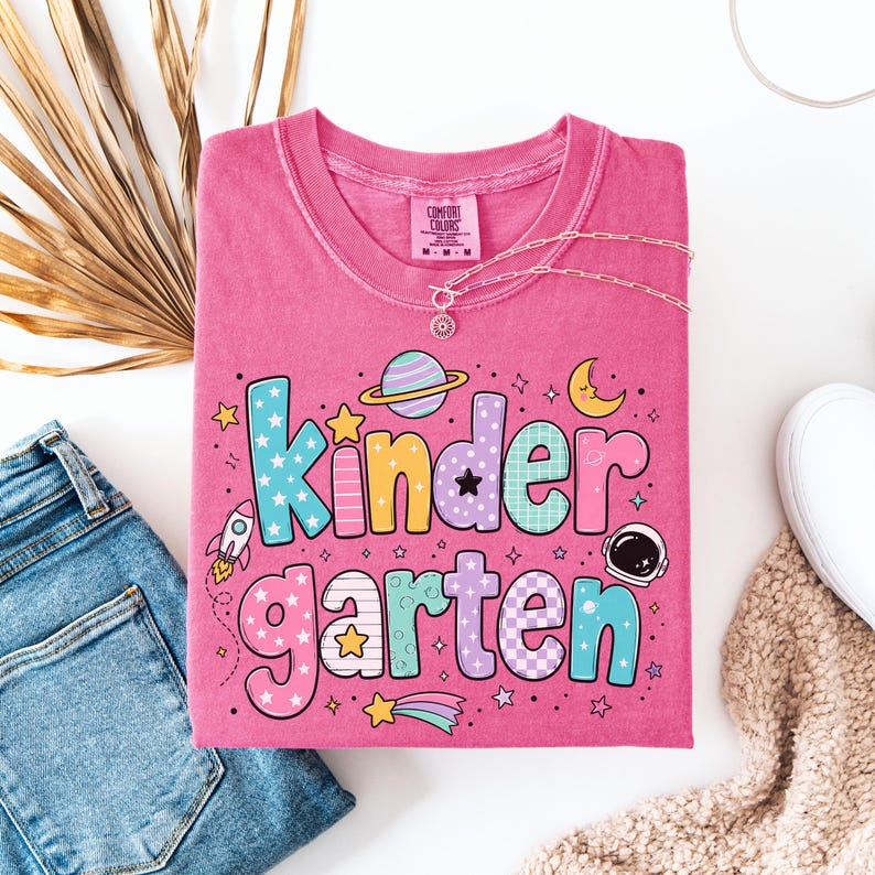 May include: A pink short-sleeve t-shirt featuring the text "Kindergarten" in large, multi-colored bubble letters. The design includes space-themed graphics such as a rocket, a planet with rings, an astronaut helmet, stars, and a crescent moon.