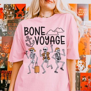 May include: Pink t-shirt with the phrase "BONE VOYAGE" and a graphic of three skeleton figures. One skeleton holds a cocktail, another a suitcase, and the third a camera. The shirt has a crew neck and short sleeves.