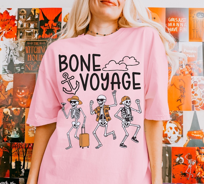 May include: Pink t-shirt with the phrase "BONE VOYAGE" and a graphic of three skeleton figures. One skeleton holds a cocktail, another a suitcase, and the third a camera. The shirt has a crew neck and short sleeves.
