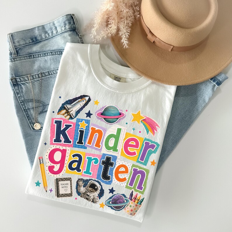 May include: A white t-shirt featuring the word Kindergarten in colorful, block-style letters. The design includes space-themed graphics like a rocket ship, planets, an astronaut, and school supplies such as a yellow pencil and a notebook labeled Teacher Notes.