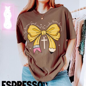 May include: A brown t-shirt with a yellow bow and a pencil with a cross on it. The text on the shirt reads "To know God and to make him known."