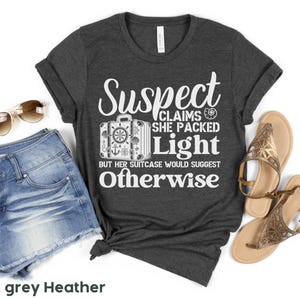 May include: Dark grey heather t-shirt with white text that reads "Suspect Claims She Packed Light But Her Suitcase Would Suggest Otherwise". The text is surrounded by a graphic of a suitcase with a nautical theme.
