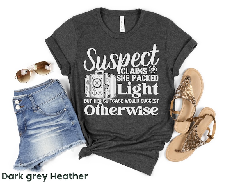 May include: Dark grey heather t-shirt with white text that reads "Suspect Claims She Packed Light But Her Suitcase Would Suggest Otherwise". The text is surrounded by a graphic of a suitcase with a nautical theme.
