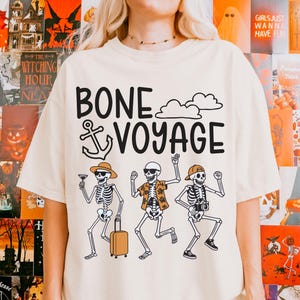 May include: Cream-colored t-shirt with the phrase "BONE VOYAGE" in black, featuring skeleton illustrations. One skeleton holds a cocktail, another a suitcase, and the third a camera. The design includes clouds and an anchor.