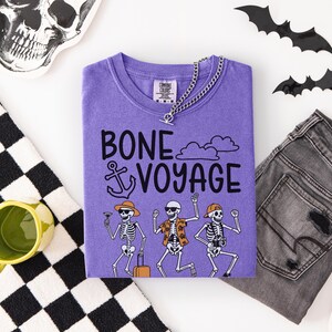 May include: A purple t-shirt with the words "BONE VOYAGE" and skeleton illustrations. The shirt is paired with gray jeans, a silver chain necklace, and Halloween decorations including bats and a skull.