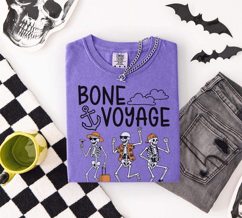 May include: A purple t-shirt with the words "BONE VOYAGE" and skeleton illustrations. The shirt is paired with gray jeans, a silver chain necklace, and Halloween decorations including bats and a skull.