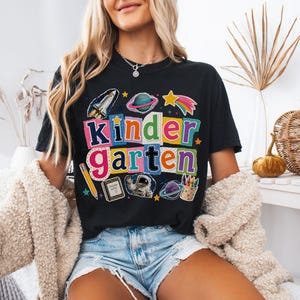 May include: A black t-shirt featuring a colorful graphic with the text "Kindergarten" in a collage style. The design includes space-themed illustrations like a rocket, planets, an astronaut, a pencil, a notebook, and a cup of crayons.