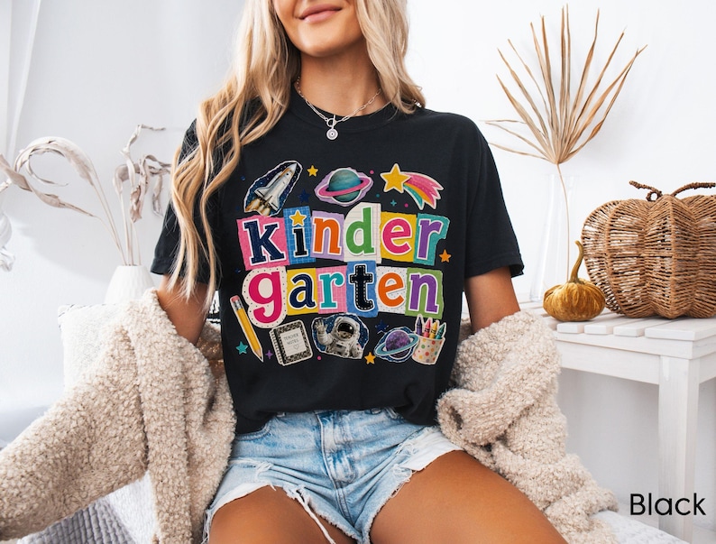 May include: A black t-shirt featuring a colorful graphic with the text "Kindergarten" in a collage style. The design includes space-themed illustrations like a rocket, planets, an astronaut, a pencil, a notebook, and a cup of crayons.