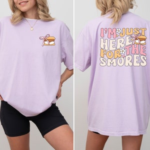 Op de afbeelding: Een lavendelkleurig T-shirt met een afbeelding van een s'more op de voorkant en de tekst "I'm just here for the smores" op de achterkant. De afbeelding heeft een roze strik en een witte bloem.