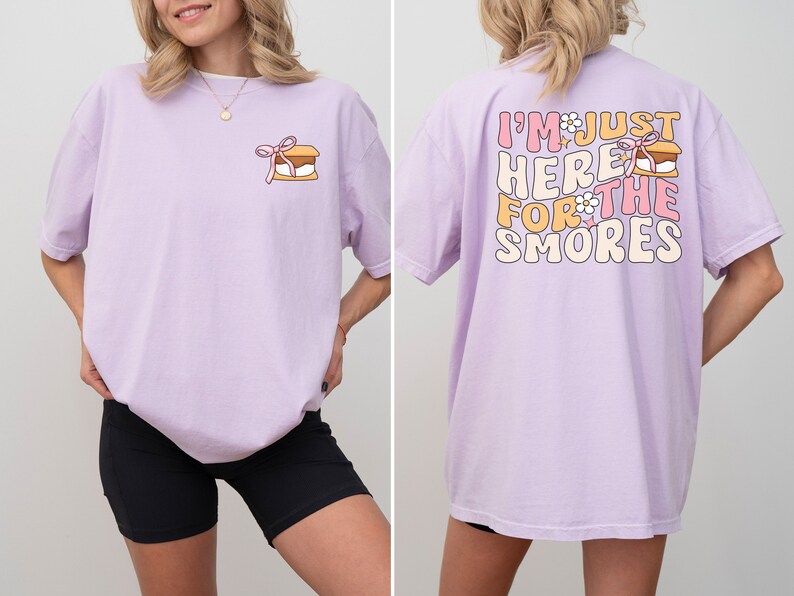 Op de afbeelding: Een lavendelkleurig T-shirt met een afbeelding van een s'more op de voorkant en de tekst "I'm just here for the smores" op de achterkant. De afbeelding heeft een roze strik en een witte bloem.