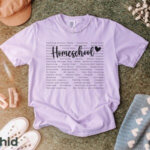 May include: A lavender t-shirt with the word "Homeschool" printed in black with a heart symbol. The shirt has a list of words related to homeschooling, including "learning without limits", "creativity", "field trips", "faith", "nature studies", "real life lessons", "individualized learning", "the world is our classroom".