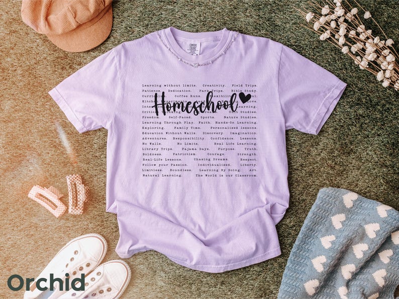 May include: A lavender t-shirt with the word "Homeschool" printed in black with a heart symbol. The shirt has a list of words related to homeschooling, including "learning without limits", "creativity", "field trips", "faith", "nature studies", "real life lessons", "individualized learning", "the world is our classroom".