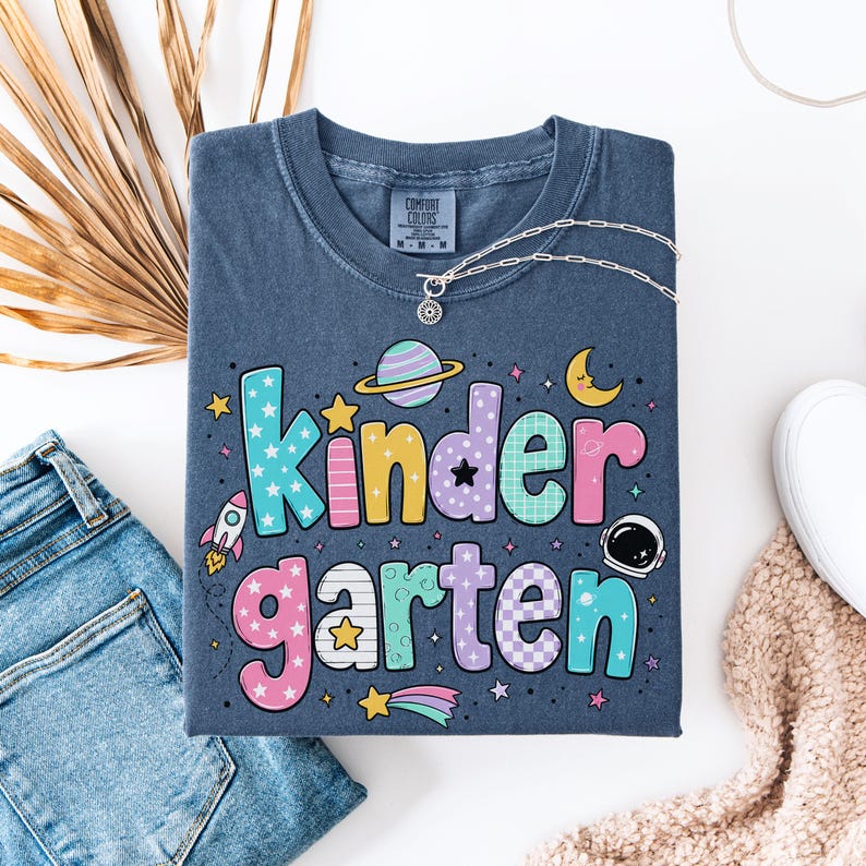 May include: A dark blue short-sleeve t-shirt with a graphic print that reads "kindergarten" in large, colorful, patterned letters. The design includes space-themed elements such as a rocket, a planet with rings, a crescent moon, and an astronaut helmet.