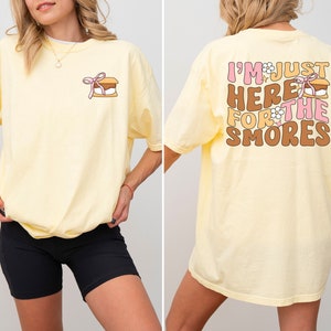 Op de afbeelding: Een geel T-shirt met een afbeelding op de achterkant die "I'm just here for the smores" zegt met een marshmallow-afbeelding. De voorkant van het shirt heeft een kleine afbeelding van een s'more.