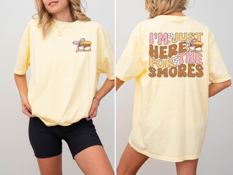 Op de afbeelding: Een geel T-shirt met een afbeelding op de achterkant die "I'm just here for the smores" zegt met een marshmallow-afbeelding. De voorkant van het shirt heeft een kleine afbeelding van een s'more.