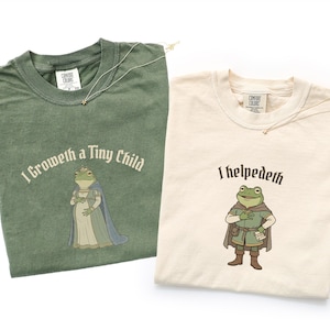May include: Two Comfort Colors t-shirts, one sage green and one cream. The green shirt has the text "I Groweth a Tiny Child" with a frog illustration. The cream shirt says "I helpedeth" with a frog illustration. Both shirts have a small gold necklace.