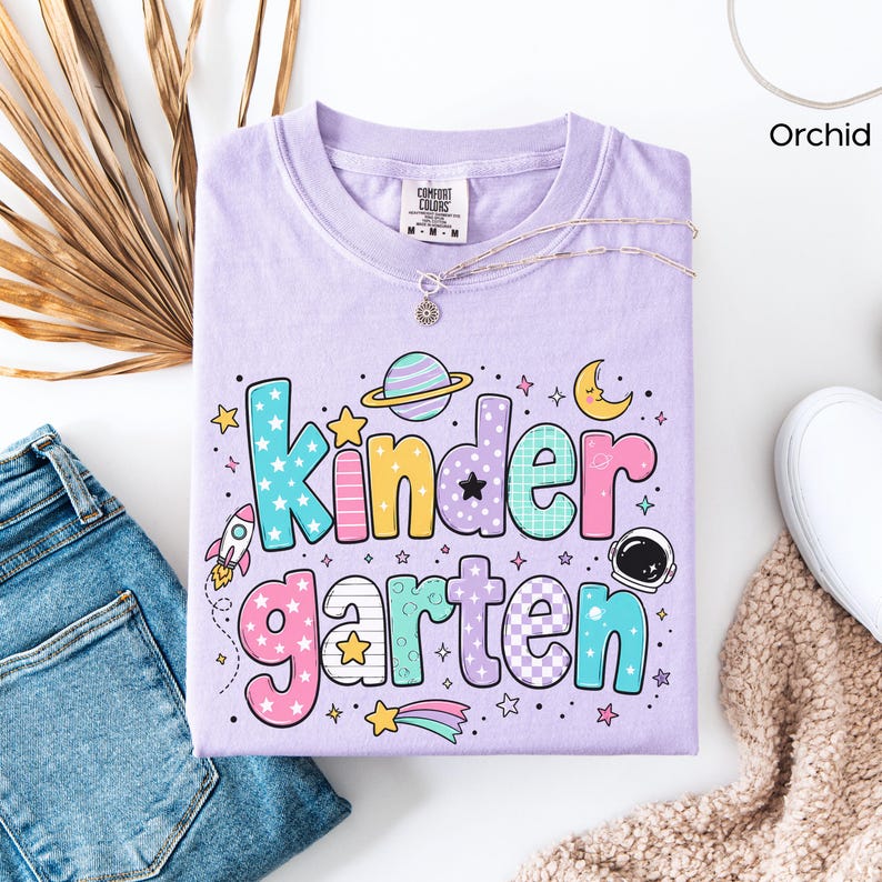 May include: An orchid-colored t-shirt featuring the word Kindergarten in large, multi-colored, patterned bubble letters. The design includes space-themed graphics such as a planet with rings, a rocket ship, an astronaut helmet, and stars.