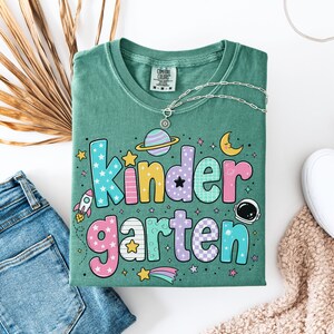 May include: A sage green t-shirt featuring the word Kindergarten in large, multi-colored, patterned bubble letters. The design includes space-themed graphics such as a rocket, a planet with rings, a crescent moon, and an astronaut helmet.