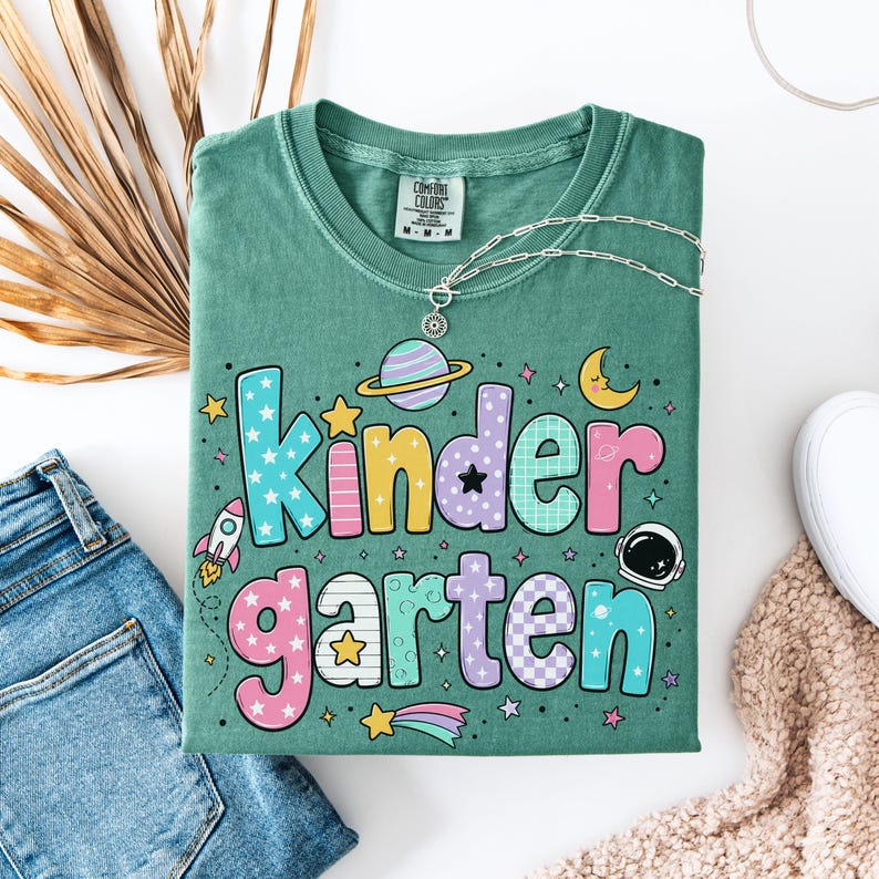 May include: A sage green t-shirt featuring the word Kindergarten in large, multi-colored, patterned bubble letters. The design includes space-themed graphics such as a rocket, a planet with rings, a crescent moon, and an astronaut helmet.