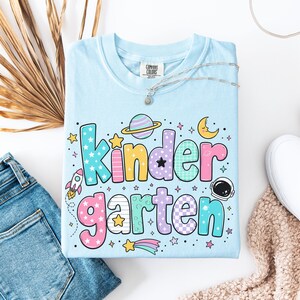 May include: A light blue t-shirt featuring the word "kindergarten" in large, colorful, patterned bubble letters. The design includes space-themed graphics such as a rocket, a planet with rings, a crescent moon, an astronaut helmet, and stars.