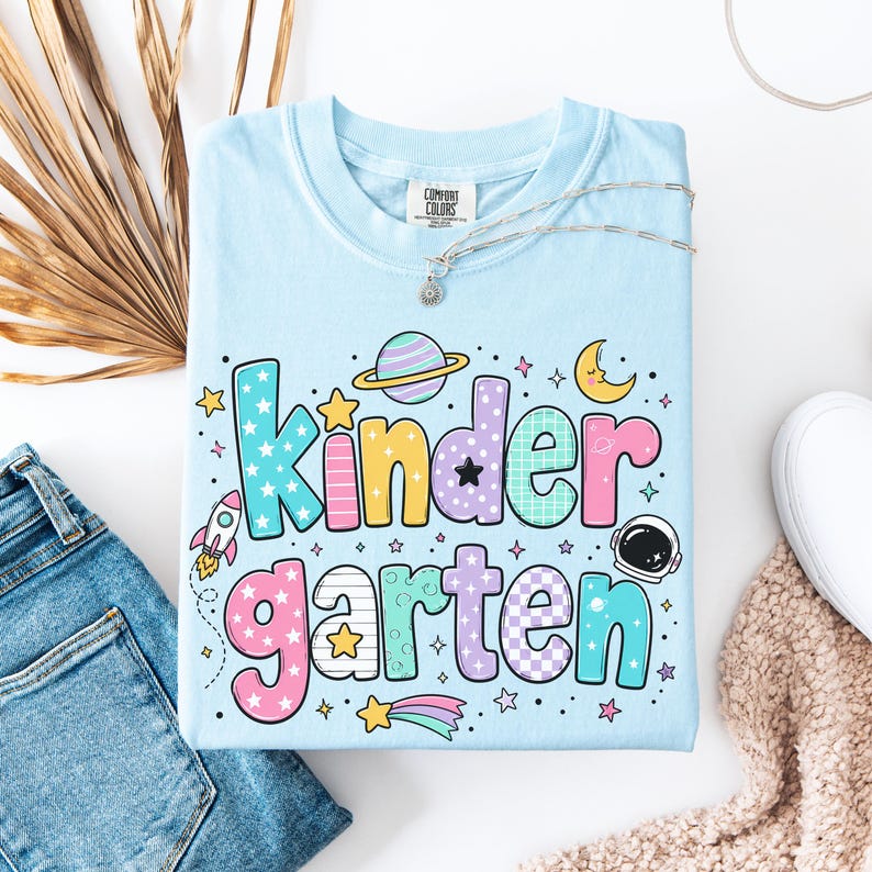 May include: A light blue t-shirt featuring the word "kindergarten" in large, colorful, patterned bubble letters. The design includes space-themed graphics such as a rocket, a planet with rings, a crescent moon, an astronaut helmet, and stars.
