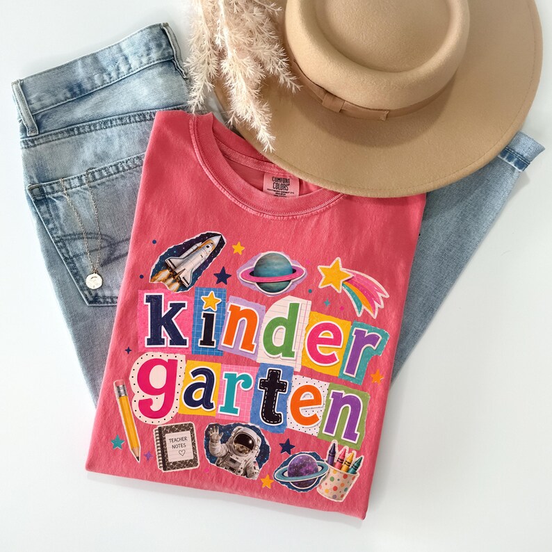 May include: A coral pink t-shirt featuring the word Kindergarten in colorful, collage-style letters. The graphic includes a space shuttle, planets, a shooting star, an astronaut, a pencil, a notebook labeled Teacher Notes, and a cup of crayons.