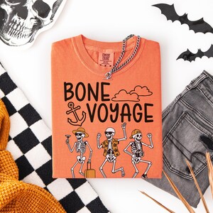 May include: Orange t-shirt with the phrase "BONE VOYAGE" and skeleton illustrations. The shirt is paired with gray jeans, a silver chain necklace, and Halloween decorations. A black and white checkered scarf and a yellow sweater are also in the image.