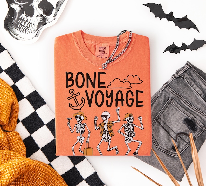 May include: Orange t-shirt with the phrase "BONE VOYAGE" and skeleton illustrations. The shirt is paired with gray jeans, a silver chain necklace, and Halloween decorations. A black and white checkered scarf and a yellow sweater are also in the image.