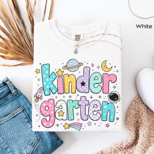 May include: A white t-shirt featuring the text Kindergarten in large, colorful, patterned bubble letters. The design includes space-themed graphics such as a rocket, a planet with rings, a crescent moon, an astronaut helmet, and various stars.