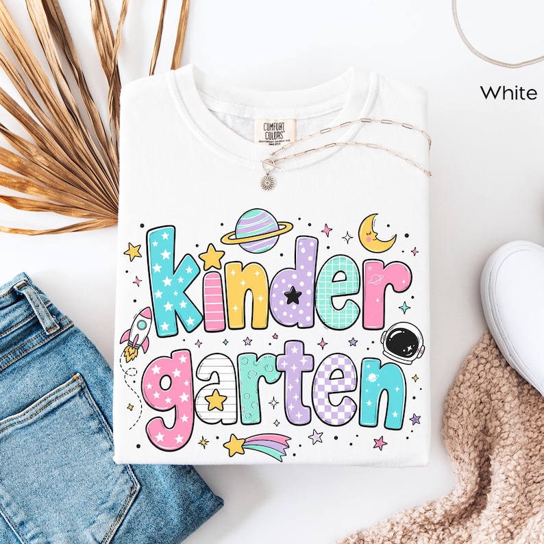 May include: A white t-shirt featuring the text Kindergarten in large, colorful, patterned bubble letters. The design includes space-themed graphics such as a rocket, a planet with rings, a crescent moon, an astronaut helmet, and various stars.