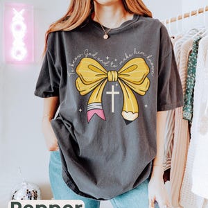 May include: A dark gray t-shirt with a yellow pencil bow and a cross. The text on the shirt reads "To know God and to make him known."