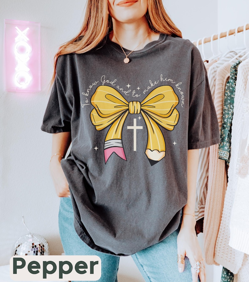 May include: A dark gray t-shirt with a yellow pencil bow and a cross. The text on the shirt reads "To know God and to make him known."