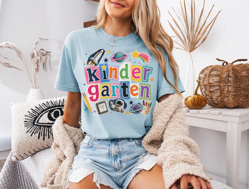 May include: A light blue t-shirt featuring the word kindergarten in colorful, blocky letters. The design includes space-themed graphics like a rocket, planets, and stars, alongside school supplies such as a pencil, a notebook, and crayons.
