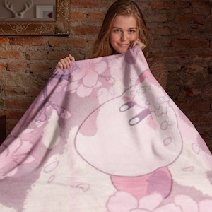 May include: A pink and white fleece blanket with a floral pattern and a cartoon character in the center. The character is white with pink accents and has two small arms.
