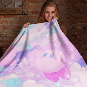 May include: A pink and purple fleece blanket with a cartoon character of a pink creature with large eyes and a pink bow. The blanket has a white background with clouds and stars.