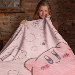 May include: A pink fleece blanket with a cartoon character design featuring a pink creature with large eyes and a small mouth. The blanket has a white background with clouds and other cartoon characters.
