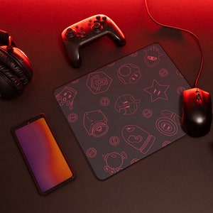 May include: A black gaming mouse pad with a pink outline of various video game characters and symbols. A black wireless controller, a black headset, a black smartphone, and a black gaming mouse are also on the surface.